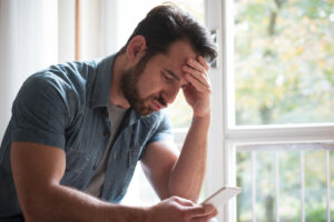 a person puts their hand on their forehead while sitting down and looking at a mobile phone in a concerned manner